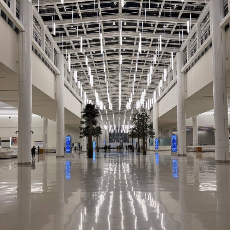 A spacious, modern airport terminal with a shiny, reflective floor. The high ceiling is adorned with a grid of vertical, LED lights hanging in rows, creating a bright, open atmosphere. Tall columns are placed along the sides, and there are several trees evenly spaced in the center of the terminal, adding a touch of greenery. Digital information boards with blue