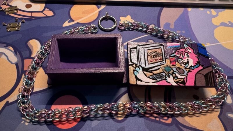 A colorful scene features a purple rectangular box, with its lid depicting a cartoon anthropomorphic animal using a computer. The character is smiling, sitting at a desk with a keyboard, mouse, and a drink. A multicolored chain made of interconnected rings is attached to the box. The background is a playful, abstract design with various colors and shapes,