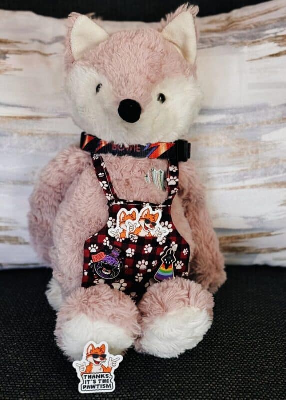 A fluffy pink stuffed animal resembling a fox is seated against a neutral background. It wears a colorful collar and a red and black checkered outfit with white paw prints. The outfit is decorated with several stickers and pins, including cartoon foxes and a rainbow paw print. A sticker at the base says, Thanks, it's the pawtism, featuring