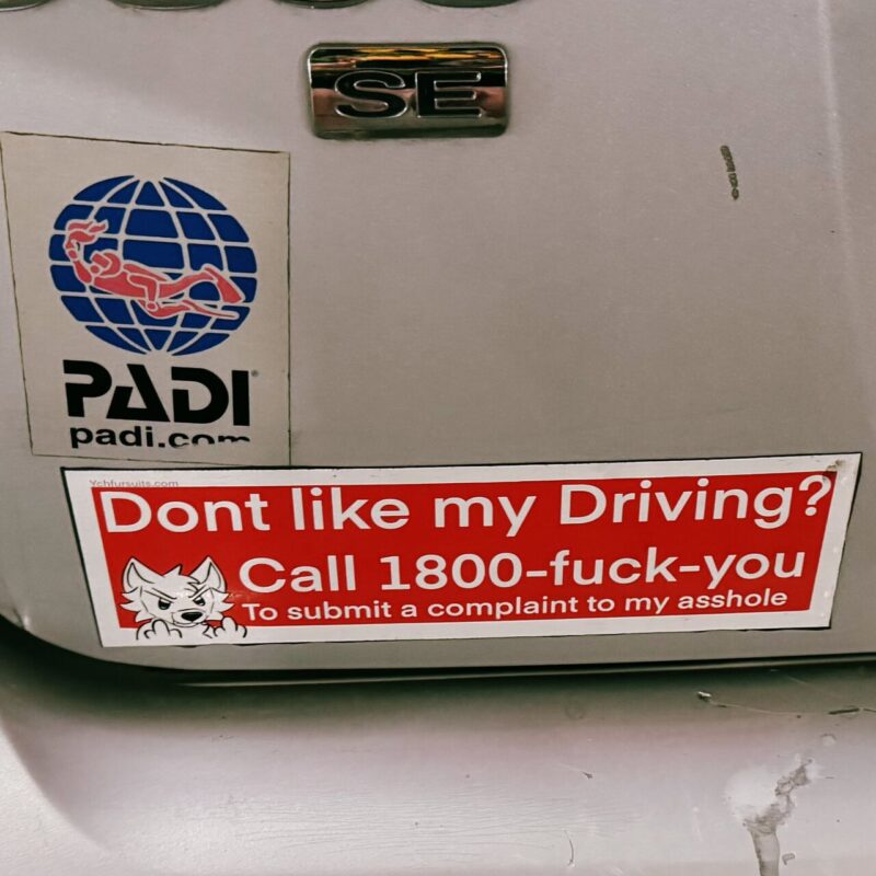 The image shows the rear of a vehicle with two stickers. The top sticker features the PADI logo, a globe with a diver, and the URL padi.com. The sticker below is a humorous red sign with white text that reads: Don't like my driving? Call 1800-fuck-you. To submit a complaint to my asshole