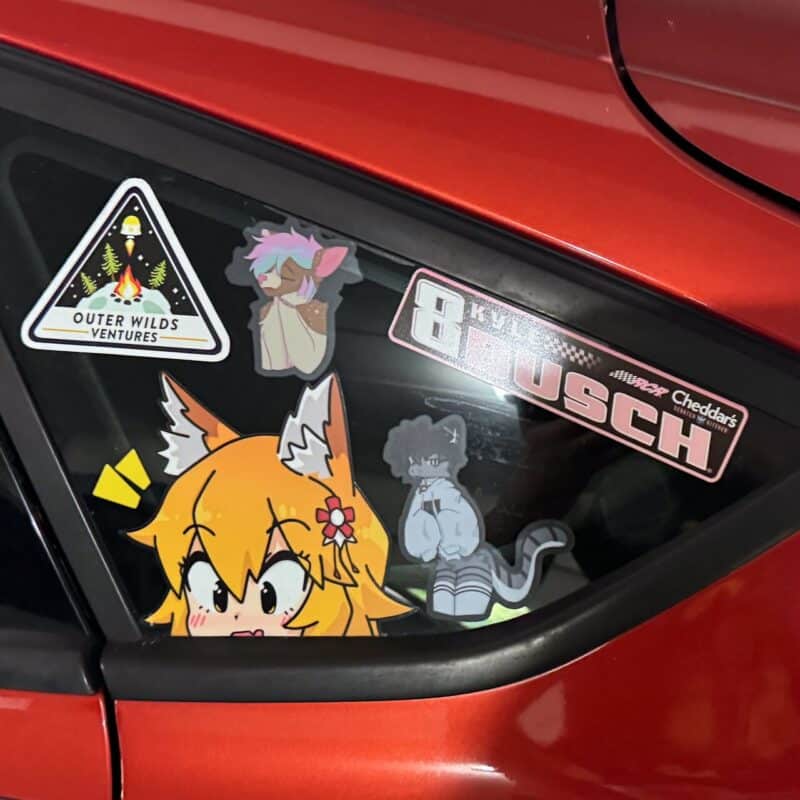 The image shows the rear window of a red car adorned with several stickers. One sticker features a cartoon character with orange hair and fox ears peeking up from the bottom. Another depicts a gray cat-like character sitting down. There is a triangular Outer Wilds Ventures sticker with stars and trees, and a NASCAR-themed sticker that says 8 Kyle Busch