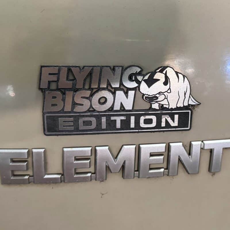 Close-up image of a car badge on a vehicle. The badge features text Flying Bison Edition alongside an illustration of a stylized, cartoonish bison marked with an arrow on its head. Below this is the word Element in larger letters. The badge and text appear metallic against a beige background. The image suggests a custom or