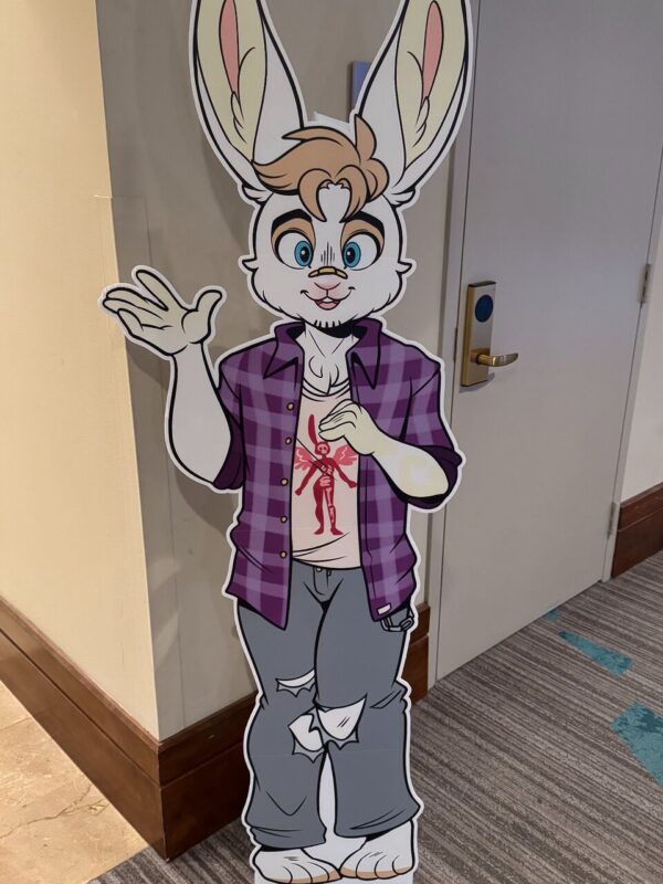 A life-sized standee of an anthropomorphic cartoon rabbit character with tall ears. The rabbit has light fur, blue eyes, and short, styled hair. It's wearing a purple flannel shirt over a white T-shirt with a red design, and gray pants with patches. The character is standing indoors, waving with its left hand. The background features a