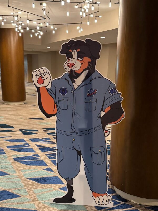 The image shows a life-size standee of a cartoon-style anthropomorphic dog character wearing a blue space-themed jumpsuit with patches of a planet and a shuttle. The dog has black and orange fur with white accents and is striking a relaxed pose with one paw raised, as if waving. The scene is set in a spacious indoor area with modern ceiling lights
