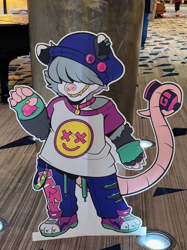 A colorful cartoon standee of a stylized anthropomorphic character with gray hair, wearing a blue hat adorned with pins, a white shirt featuring a smiley face with crossed-out eyes, and purple pants with pink accents. The character has a pink rat-like tail, green gloves, and multicolored sneakers. They are depicted standing in a room with