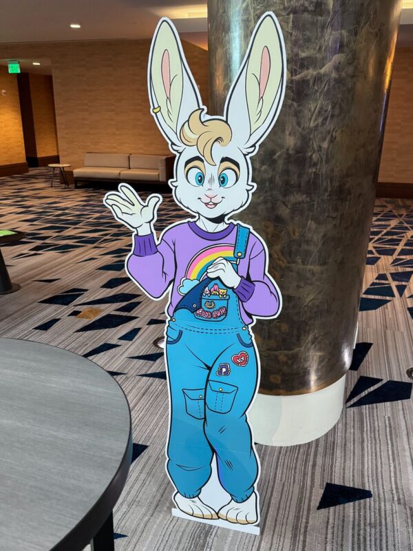 A life-sized cardboard cutout of an anthropomorphic rabbit with large ears, a tuft of blond hair, and blue eyes stands in a hotel lobby. The rabbit is cartoonish, wearing a purple sweatshirt with a rainbow design and blue overalls featuring playful patches, including a music note and a heart. It has a friendly expression and is waving with one
