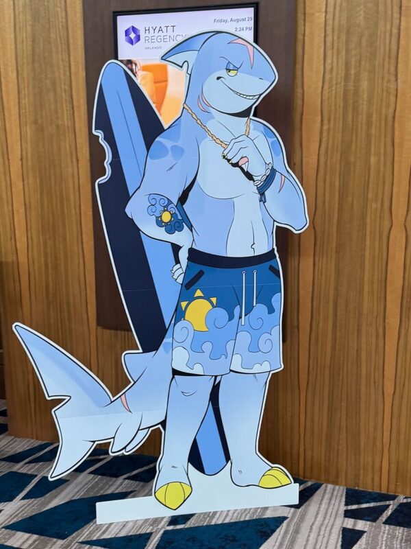 A life-sized cardboard cutout of an anthropomorphic shark character is positioned indoors, against a wooden wall backdrop. The shark is light blue, muscular, and depicted with a sly smile. It wears sunglasses, a gold chain, and beach shorts with a sun and wave pattern. Yellow flip-flops complete the look. Behind the cutout, a sign