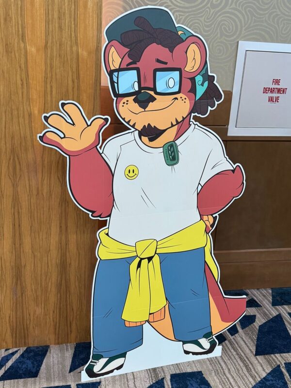 A life-sized cardboard cutout of a cartoon anthropomorphic character stands against a wooden panel. The character has red fur, wearing glasses and a backward cap. It smiles and waves with one hand. Dressed in a white t-shirt with a smiley face, blue pants, and green sneakers, it has a yellow sweatshirt tied around its waist. A