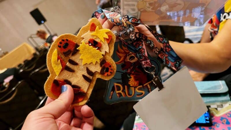 A hand is holding a felt tiger ornament with bright colors and intricate details. In the background, another person holds a colorful lanyard featuring a badge with an illustrated character and the text Rust. The setting appears to be a conference or event with chairs and people in the blurred background. The image conveys a playful and creative atmosphere.