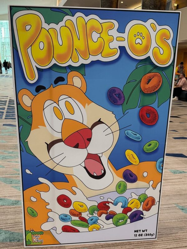 A colorful, cartoon-style poster depicts a cheerful tiger above a bowl of cereal with vibrant, multicolored rings. Splashes of milk surround the cereal. The word “POUNCE-O’S” is boldly displayed at the top in yellow and white, with a paw print in the letter O. Tropical leaves fill the background. The bottom left