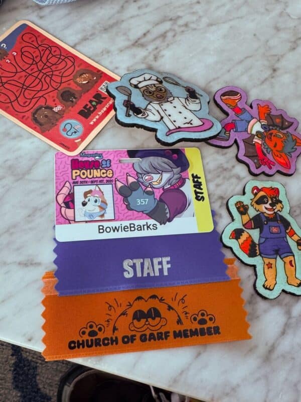 A collection of themed convention items is displayed on a marble surface. There are various colorful patches featuring cartoon animal characters, such as a raccoon in a chef outfit holding utensils, and another character in vibrant, playful poses. A red card shows a maze with illustrations of bears. A convention badge with the name BowieBarks titled House