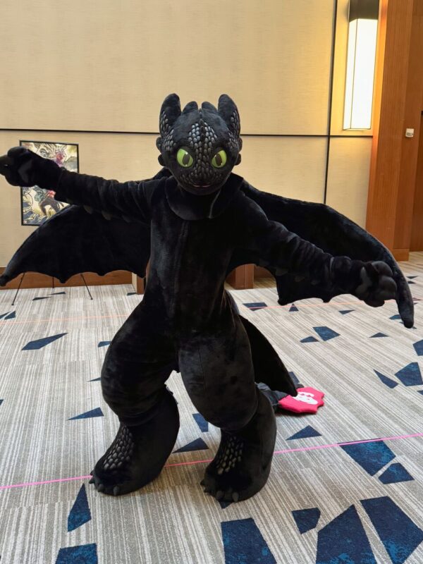 A person in a Toothless dragon costume from How to Train Your Dragon stands indoors on a patterned carpet. The costume is black with large green eyes and outstretched wings. The dragon's body is detailed with textured scales. In the background, the beige walls feature a framed artwork. The lighting is warm, coming from a modern wall lamp