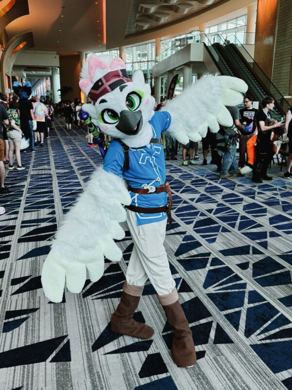 A person in a white and blue bird costume poses playfully in a convention center hallway. The costume has large wings, green eyes, and a pink accent on the head. The character wears a belt and brown boots with a patterned floor beneath. People in casual clothing walk or stand in the background, and escalators are visible on the right, with