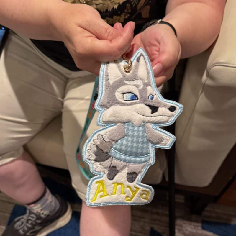 A person is holding a large, embroidered keychain featuring a cartoon-style wolf character. The wolf has light gray fur, a white face, and is wearing a blue sweater. The character appears to be smiling and has a friendly expression. Below the wolf, the name Anya is embroidered in yellow letters. The person's hands are visible, and