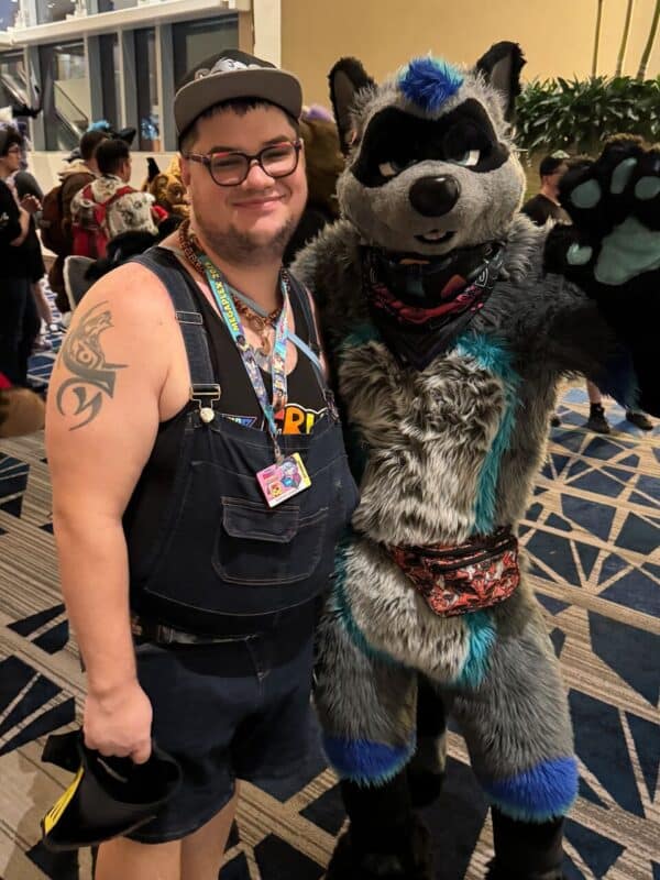 A person stands smiling next to an individual in a gray and blue wolf fursuit with black accents. The person is wearing a cap, black tank top, and overalls, with a tattoo visible on their left arm. They have a lanyard around their neck and are holding a small item in their hand. The fursuited character has