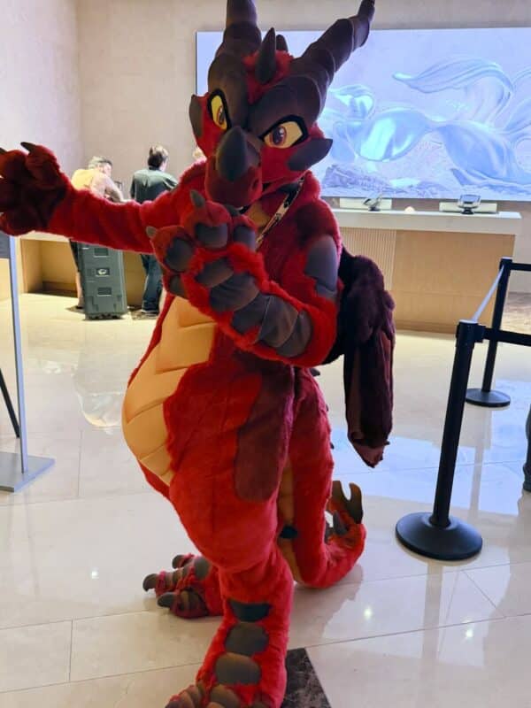 A person is wearing a colorful dragon costume with vibrant red and brown features. The costume has large eyes, protruding horns, and a textured tail. The person is posing energetically in an indoor setting with a marble floor. Behind them, a modern reception area is visible, featuring a glowing abstract art installation on the wall. The atmosphere is lively,