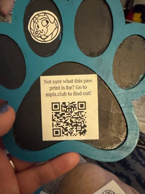 A blue and black wooden paw print features a small sticker of a cartoon dog's face in the upper section. Below, a label reads, Not sure what this paw print is for? Go to mplx.club to find out! accompanied by a QR code. A hand with blue-painted nails holds the paw print at the bottom.
