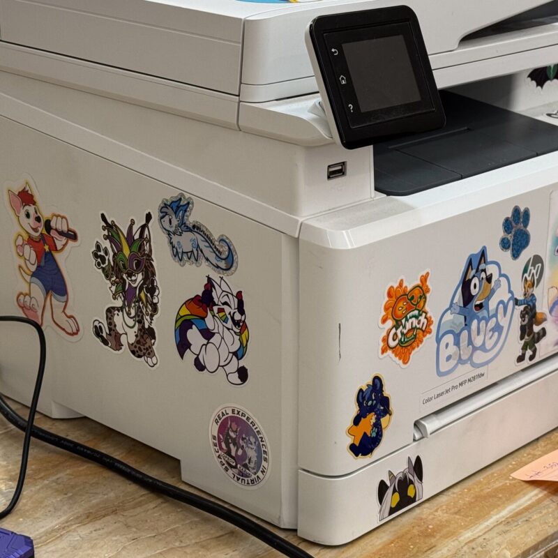 A white printer adorned with colorful stickers featuring various cartoon and animal characters. The stickers include drawings of anthropomorphic creatures, such as a playful fox, a rainbow-striped dog, a dragon, and a wolf. One sticker has the text Bluey with a cartoon wolf, while another features a paw print. The printer has a small touchscreen display and