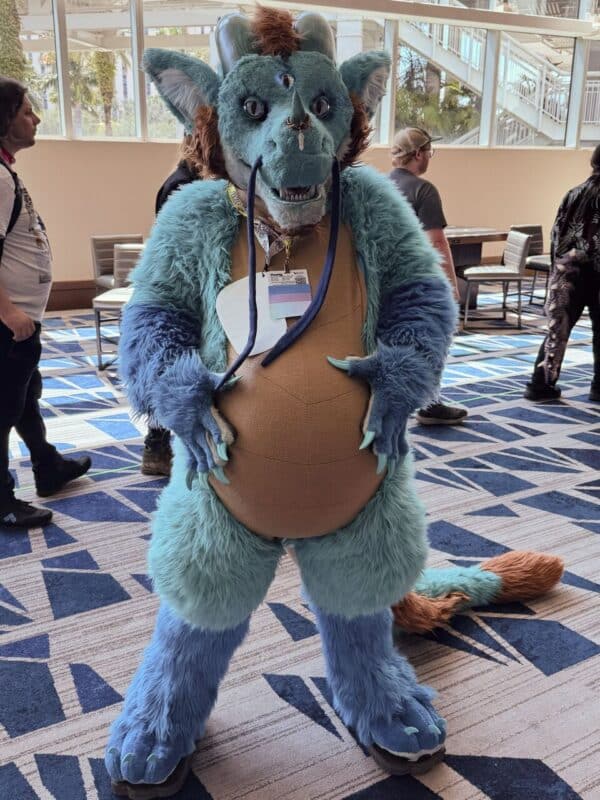 A person is wearing a furry costume, resembling a blue and brown creature with large ears, horns, and a long tail. The costume features a round belly, claws, and a third eye on the forehead. The person is standing indoors on a geometric-patterned carpet with natural light coming from large windows in the background. Other people are casually walking and