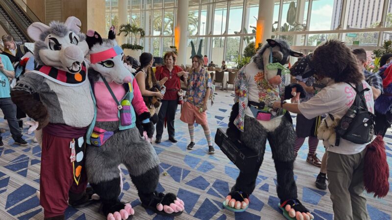 A lively indoor scene at a convention featuring people in colorful animal costumes, including a large mouse and a wolf with accessories like a headband and glasses. The background shows a spacious lobby with attendees dressed in various costumes, conversing and enjoying the event. The floor is patterned with blue geometric designs, and large windows let in natural light. There are plants