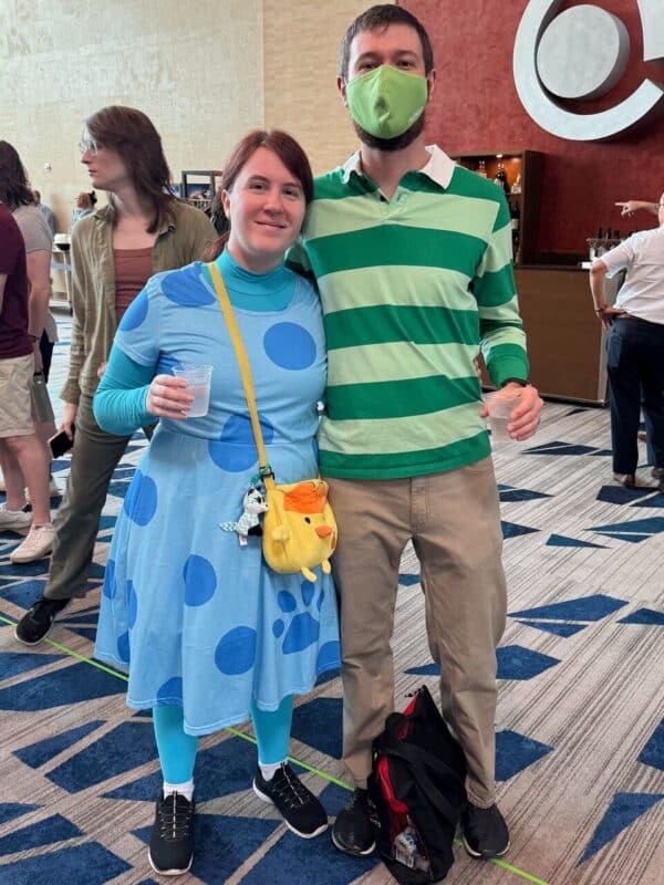A couple in colorful costumes stands together in a large indoor setting. The woman wears a blue dress with large blue spots and a bright yellow bag shaped like a bird. She holds a drink in one hand. The man wears a green striped shirt, beige pants, and a green mask. They both appear to be attending an event, surrounded by other people