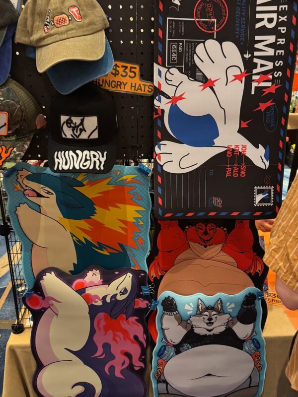 A display of various themed hats and pillow designs is shown. The hats feature logos and the word Hungry, alongside embroidered images of food like steak and sushi. The pillows, hung vertically, display cartoonish designs of animals and creatures, including a fiery dinosaur-like character and a smiling, anthropomorphic wolf. A sign indicates the hats are priced at