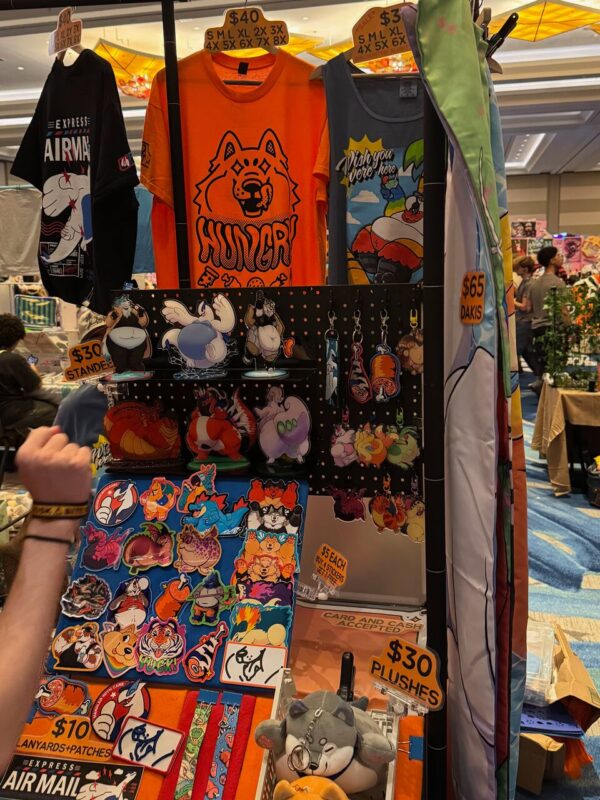 A vibrant convention stall displays various merchandise. Prominently featured are colorful T-shirts, including an orange one with a cartoon dog and the word HUNGRY. Alongside are tank tops and an assortment of plush toys priced at $30. A section showcases stickers, pins, and lanyards with cartoon characters. Prices range from $5