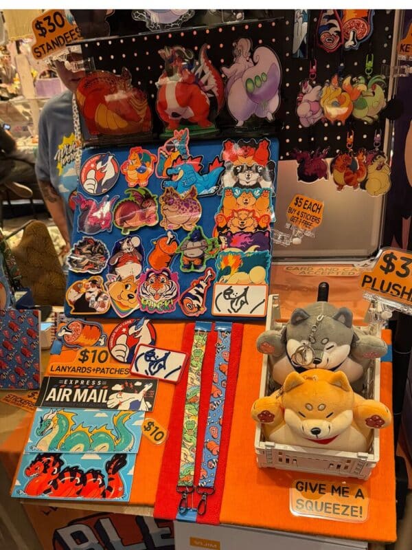A colorful display at a vendor booth featuring various cartoon-themed merchandise. There are acrylic standees, vibrant stickers, keychains, and plush toys. The standees and stickers depict playful animal characters in imaginative designs. A sign lists prices: Standees $30, Stickers $5 each or 4 for $15, and L