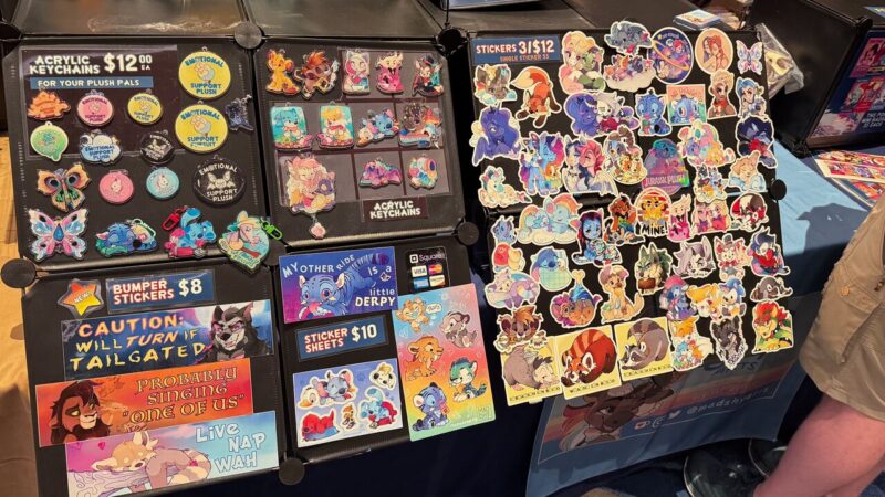 A display features various colorful items for sale, including acrylic keychains, stickers, and bumper stickers. The keychains depict cartoonish animals and characters, priced at $12 each. To the right, a board displays an assortment of vibrant stickers with different characters and designs, priced at 3 for $12 or $5 each. Below, bumper stickers