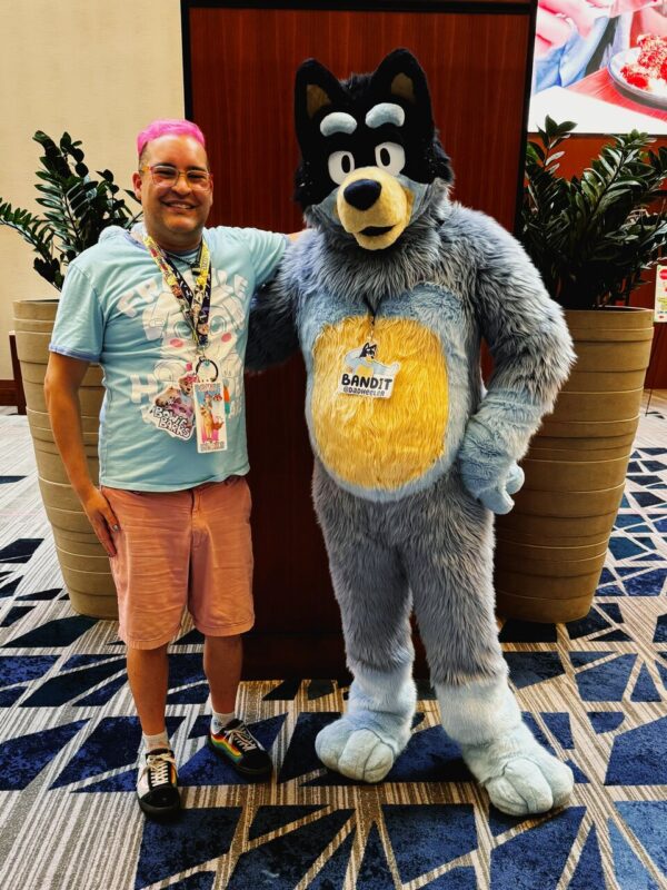 A person with pink hair is smiling while standing next to someone in a large, furry costume resembling a cartoon dog, with blue and gray fur and a yellow belly. A name tag reads Bandit on the costume. The person is wearing a light blue shirt, pink shorts, and rainbow sneakers. They are indoors, with patterned carpet and large
