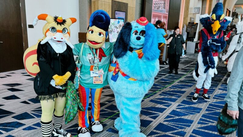 A group of people in colorful furry costumes stands together in a convention hallway. The first person wears a tan and orange-themed cat suit with large eyes and striped tail. The second person is dressed as a character in a blue and red outfit with an exaggerated headpiece. The third costume is a fluffy blue dog with black floppy ears and a red nose.