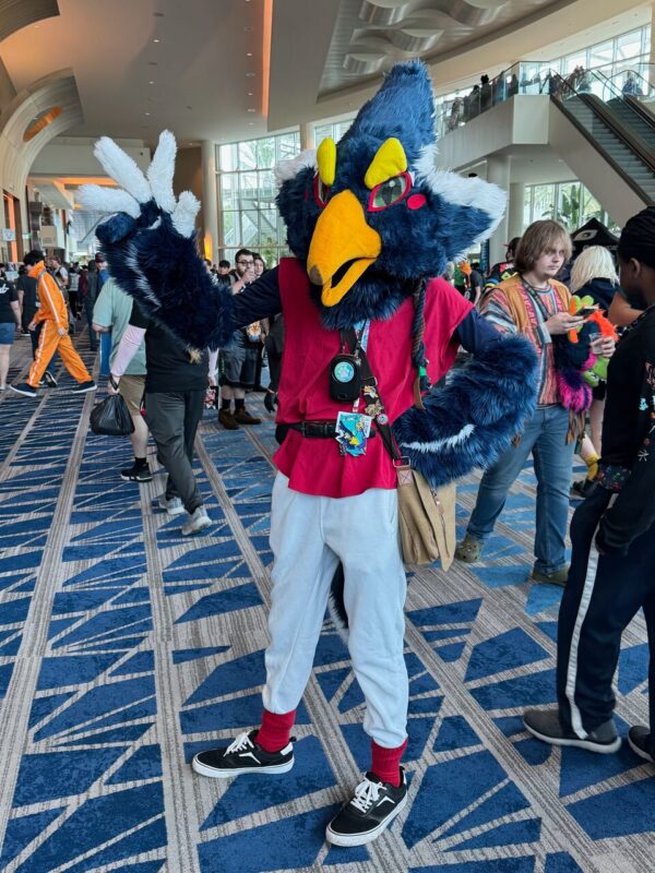 A person is cosplaying in a bird-like costume in a convention center. The costume features blue feathers, a large yellow beak, and red accents around the eyes, paired with a red vest and white pants. The individual is posing confidently, with one hand making a peace sign. Other attendees in casual attire are visible in the background, along with
