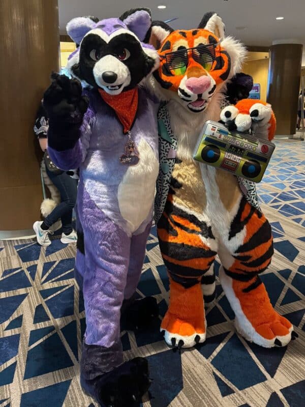 Two people in animal costumes pose for a photo. The one on the left wears a purple and white raccoon suit with a red bandana and a necklace. The one on the right is dressed in an orange and black tiger costume with sunglasses and a patterned shirt, holding an inflatable boombox. They are standing on a carpet with a geometric pattern