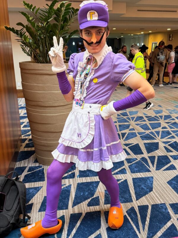 A person is wearing a cosplay outfit inspired by a video game character. The costume includes a purple maid dress with white frills, purple gloves, and tights. They wear orange clogs and a purple cap with a logo. The person poses with one hand on their hip and the other making a peace sign. They have a painted-on mustache and