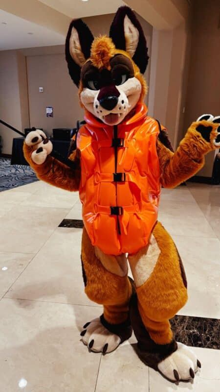 A person in a detailed furry costume resembling a canine stands in a hotel lobby. The costume is predominantly brown with white and black accents, featuring large, expressive eyes and a playful expression. The person wears a bright orange, shiny life vest, giving the costume a whimsical touch. Their paws are up, one holding a black leash or strap, and they