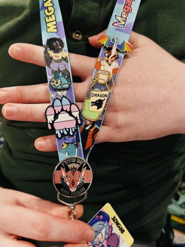 A person is holding a colorful lanyard with various decorative pins and badges. The lanyard has illustrations, including one of a person with dark hair and vibrant attire. There is a dragon-themed pin with a rainbow umbrella. Another pin displays a stylized paw print in pastel colors. A round badge shows an animal face with Certified Scum