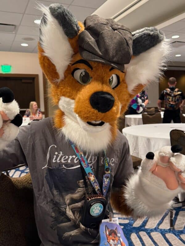 A person is wearing an orange and white fox costume, complete with a fuzzy hat and large ears. The costume includes detailed face markings and a realistic snout. They are inside a convention hall with round tables and chairs, and other attendees are visible in the background. The person is wearing a shirt with an illustration, and a lanyard with badges