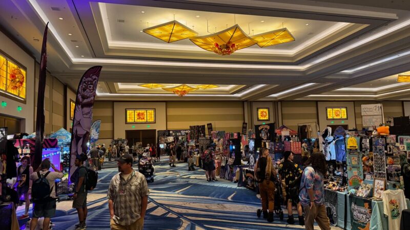 A spacious convention hall filled with attendees exploring various vendor booths lined up on both sides. Each booth displays colorful merchandise such as clothing, art prints, and plush toys. The ceiling is adorned with bright, geometric light fixtures, and decorative stained-glass windows enhance the warm lighting. People of different ages are browsing the stalls, some stopping to chat with vendors