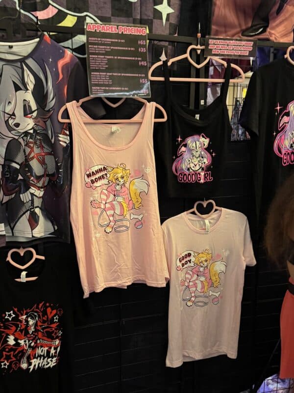 A display of graphic t-shirts on heart-shaped hangers, featuring various anime and cartoon-style designs. The top left t-shirt has a wolf character with stars. In the center, a pink tank top with a fox and Wanna Bone? text, and a black tank with a character and Good Girl text. To the right, a