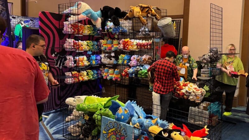 A bustling booth at a convention features a variety of colorful plush toys on display. The shelves are filled with toy animals in various colors and sizes. Three people are behind the booth: one in a floral shirt helping a customer, another in a green shirt managing inventory, and a customer wearing a red plaid shirt examining items. Bright lighting highlights the vivid colors