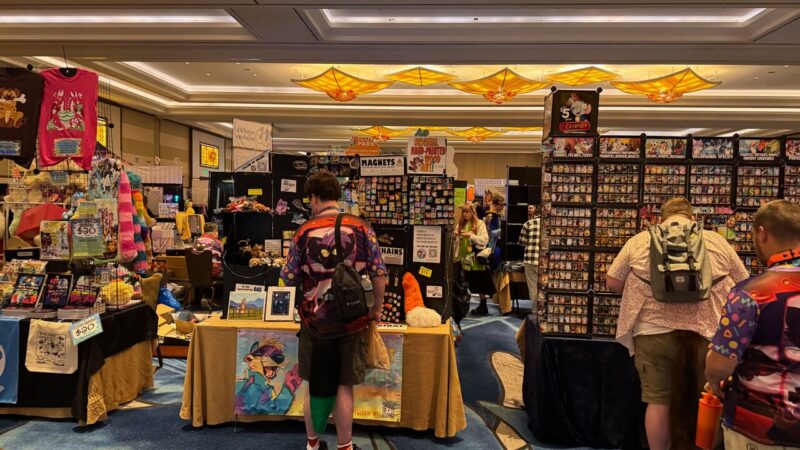 A bustling convention hall filled with colorful merchandise and attendees. Various stalls display an array of items, including T-shirts, plush toys, framed artwork, and magnets. The ceiling is adorned with yellow, geometric light fixtures. People are browsing the booths, with some wearing vibrant shirts and carrying backpacks. Signage is visible, advertising prices and products like hand-painted