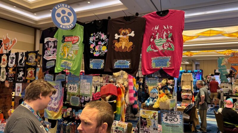 A colorful vendor booth at a convention displays a variety of graphic T-shirts hanging prominently. Designs include cartoon creatures and playful text. Below the shirts are price lists showing prices for T-shirts, tank tops, and plushies. Several plush toys are visible on shelves. The booth is bustling with attendees, and a Raizap logo featuring a paw