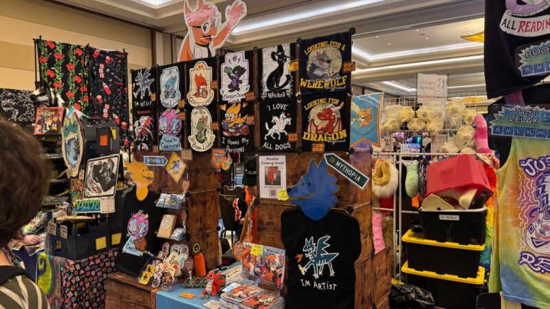 A vibrant vendor booth at a convention displays various items with colorful designs. The booth features T-shirts and banners with playful and fantasy-themed graphics, such as dragons and werewolves, each with quirky phrases. A section displays plush toys, with a sign indicating more to come. The booth is set against a backdrop of poppy-patterned fabric, and shelves