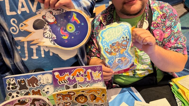 Two people display colorful, cartoon-themed artwork and sticker sheets at a table. The left person holds a circular piece featuring a panda with the word SOT in rainbow colors. The right person showcases a design featuring a playful, anthropomorphic dog with the word CHU. Below, various sticker sheets display cartoon animals such as pandas, foxes