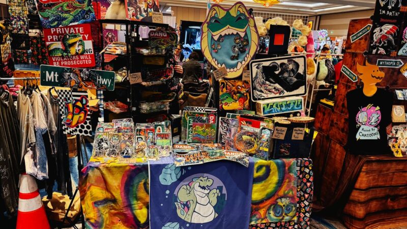 A vibrant vendor booth displays eclectic artwork and merchandise. The booth features colorful signs, apparel, and art prints with cartoonish designs, including dinosaurs and abstract patterns. A prominent sign reads Raised by Cartoons. Various items are stacked on a table covered by a bright, multicolored cloth. T-shirts hang on the left while a wooden shelf on