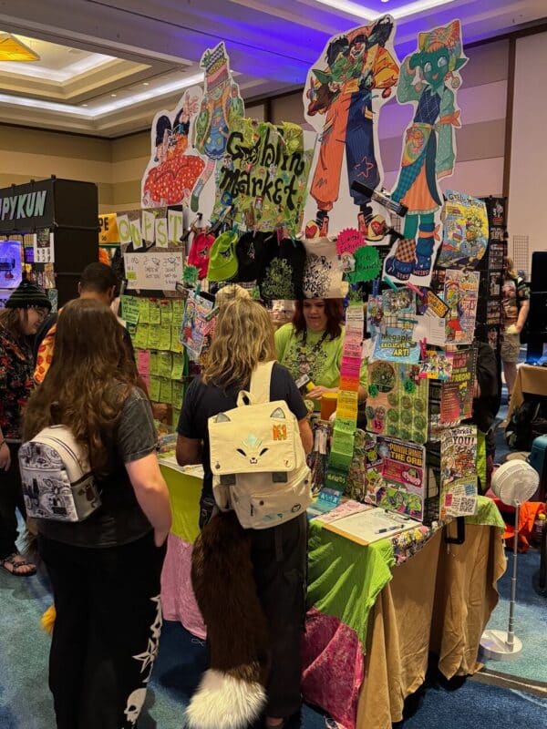 A colorful vendor booth with a sign reading Goblin Market features vibrant, cartoon-style cutouts of characters above it. The stand is adorned with green and pink cloths, offering a variety of small items and artwork. Two people with themed backpacks, one resembling a cat with a mock tail, are browsing the merchandise. The booth displays numerous eye