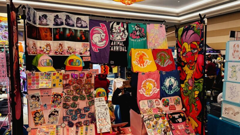 A colorful convention booth displays various merchandise, including T-shirts with playful animal graphics and phrases like Animal Cannibal and Funky Critter. Vibrant posters and banners featuring cartoon animals adorn the backdrop. A variety of colorful hats, keychains, and sticker sets with similar themes are neatly arranged on the table. The overall aesthetic is lively and