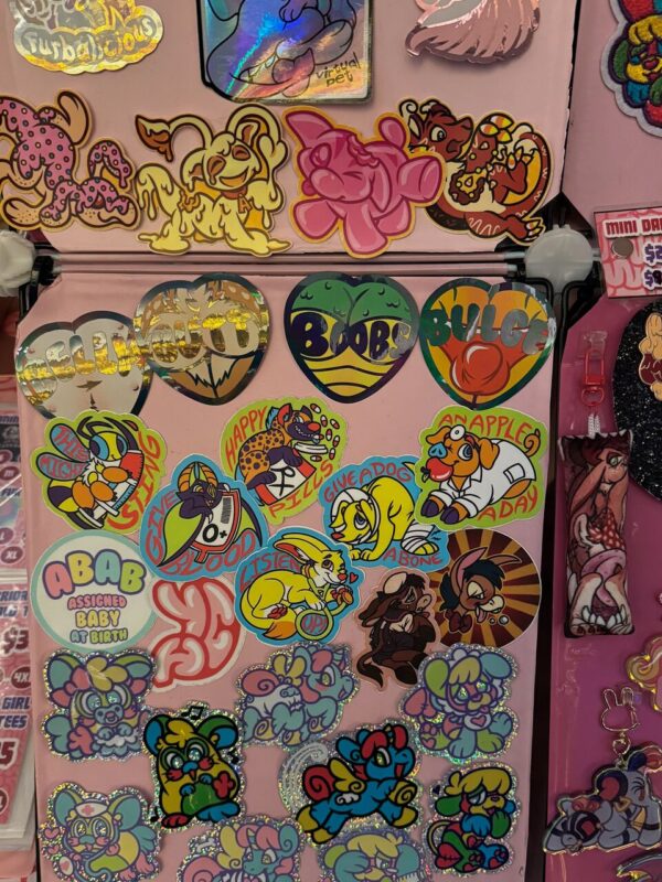 The image displays a collection of colorful stickers on a pink surface. The stickers feature various cartoonish animals and playful designs, including dog-like and rabbit-like characters in bright colors. Some stickers contain text, such as Assigned Baby at Birth and phrases like Give A Dog A Bone. The designs are vibrant, with heart-shaped stickers and shiny elements