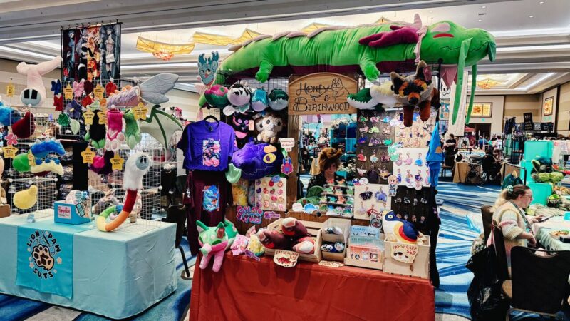 A vibrant vendor booth at a convention displays a variety of colorful plush toys, T-shirts, and handcrafted items. A large green plush dragon hangs above the booth. The table is draped with a blue cloth featuring a paw logo and is covered with plush animal tails and other soft items. The booth also features an assortment of pins, stickers, and colorful