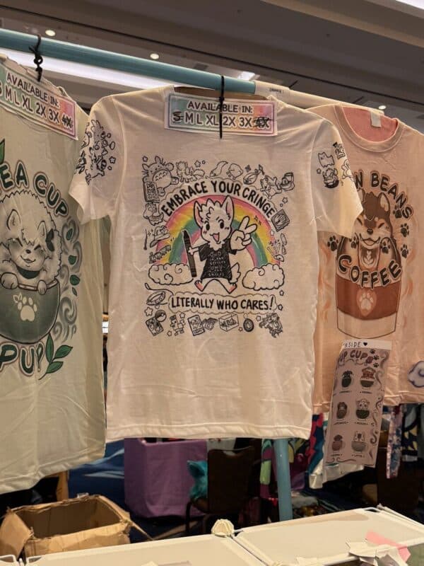 Three T-shirts hang on a rack at a convention booth. The central shirt is white and features a cartoon animal with a rainbow, surrounded by quirky doodles, with the text Embrace Your Cringe, Literally Who Cares! The shirt on the left displays a cat in a teacup, captioned Tea Cup Pup. The right